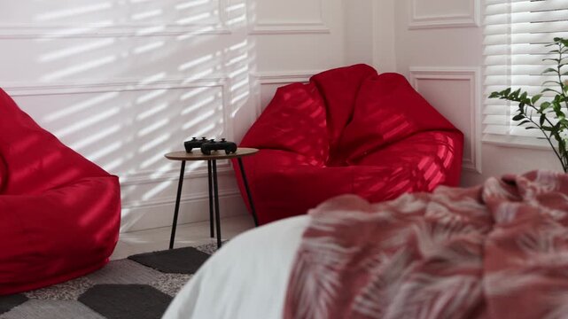 Cute little dog on bed, red bean bag chairs and wooden table with game controllers in bedroom. Camera moving left