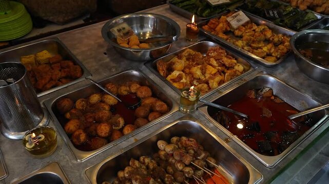 Indonesian street food buffet with assorted fried snacks, eggs, tofu, and spicy dishes in stainless trays under warm market lighting.