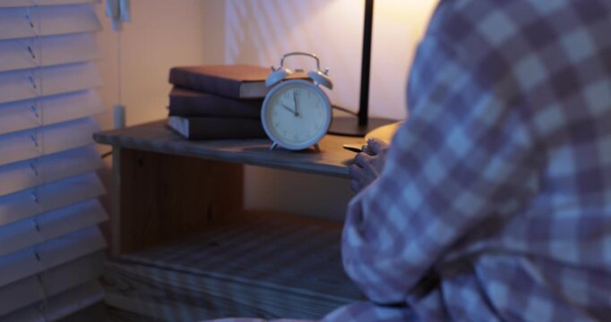 Woman setting alarm clock and putting it on bedside table indoors at night, closeup