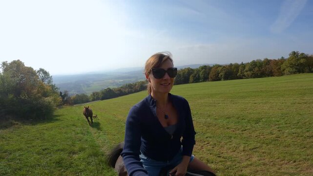 SELFIE: Smiling young woman riding a dark brown horse across sunny meadow, with a loose chestnut horse and dog following behind. Horseback ride across wide grassland with a view of hilly countryside.