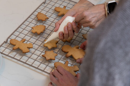 Two friends decorating gingerbread cookies on stone countertop using piping bag and cooling rack