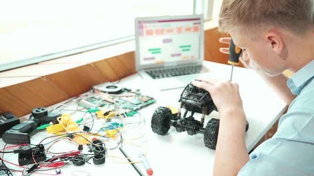 Happy teenager fixing robotic model while using electronic equipment remove car wheel with wire and laptop scatter around. Smart boy study or learn about car robot construction at class. Edification
