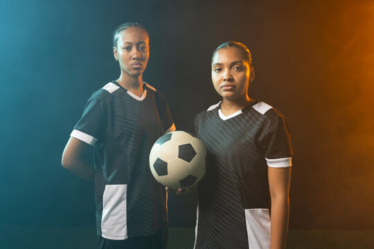 Female soccer teammates wearing matching jerseys posing side by side in studio, holding soccer ball