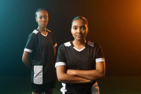 Female teammates posing on turf in studio wearing black jerseys and shorts with teal amber lights