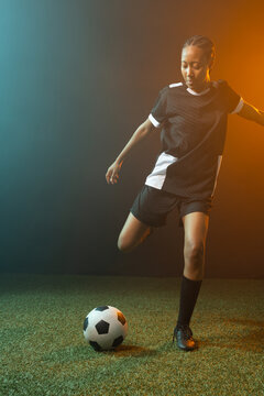 African American woman footballer balancing on left foot, black kit, ball on turf, teal light