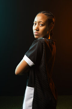 Woman posing, crossing arms, looking back, braids, jersey, teal-orange lighting on turf in studio