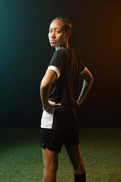 African woman soccer player posing on studio turf wearing kit and socks under teal-orange light