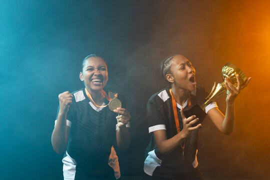 African American female teammates holding gold medal on orange ribbon and trophy in studio haze