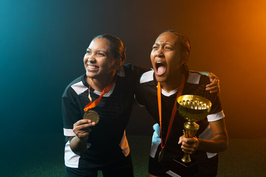 Two African American female players celebrating on turf with gold trophy and medal, lit teal or oran