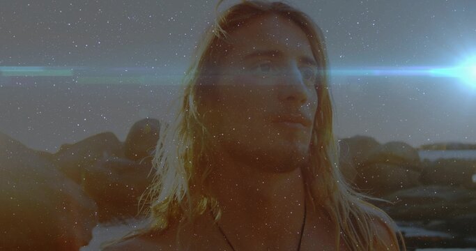 Standing shirtless man with blond hair gazing toward horizon at rocky shoreline, wearing necklace