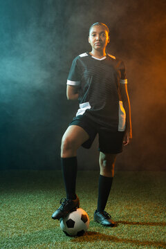 Female soccer player standing on turf wearing black-jersey, foot on ball amid teal-amber smoke