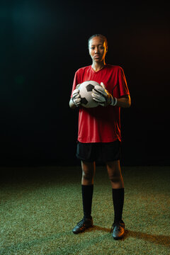 African American woman goalkeeper posing on turf holding b and w soccer ball wearing red jersey, glo
