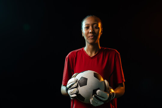 Woman GK standing in studio holding soccer ball in red jersey and gloves rim lighting blue-orange