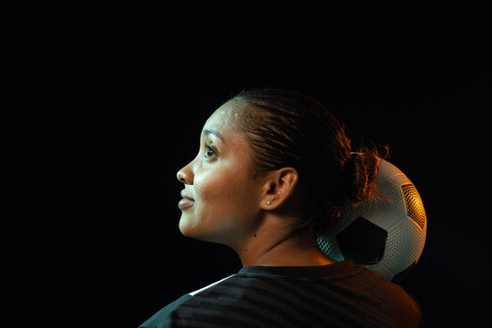 African American woman balancing soccer ball behind neck on shoulder, posing in studio with jersey