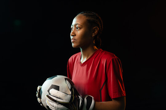 African American woman keeper in red jersey gloves studs holding soccer ball facing left in studio