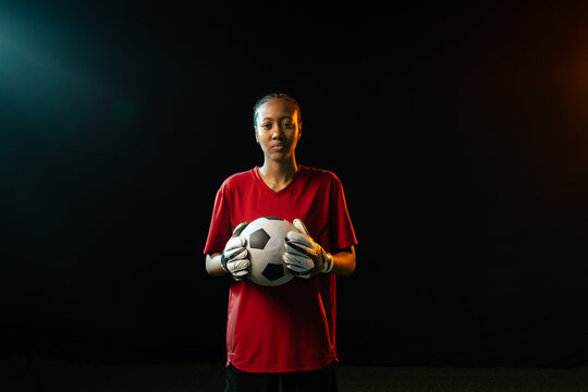 Female goalie holding soccer ball, gloved, red kit in studio under teal-orange rim lights over turf