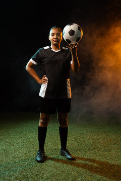 African American woman balancing soccer ball on head while posing on turf in black jersey, fog
