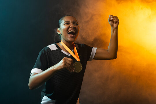 African American woman wearing jersey cheering, holding gold medal on orange ribbon in smoky studio