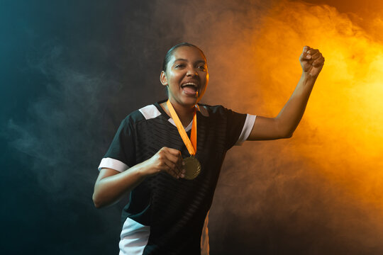 African American woman cheering holding gold medal on orange ribbon, wearing jersey in studio fog