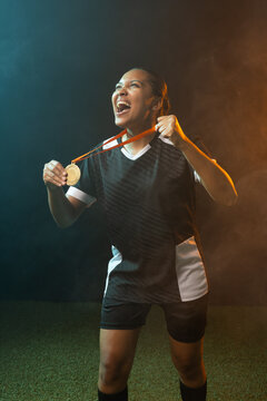 Female athlete standing on turf, holding ribboned gold medal, in black jersey, teal or orange fog