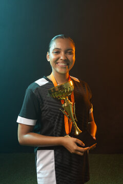 Woman wearing black jersey holding gold trophy and orange ribbon medal on studio turf