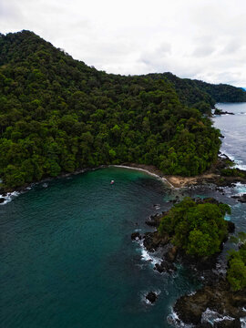Aerial view of Bahia Solano featuring dense tropical rainforest, turquoise waters, and a small boat anchored near the rocky shoreline in Mutis, Choco, Colombia.