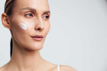 Fototapeta premium Woman applies white face cream on cheek in a close up beauty portrait, skincare and self care concept, calm expression, soft studio lighting on light background