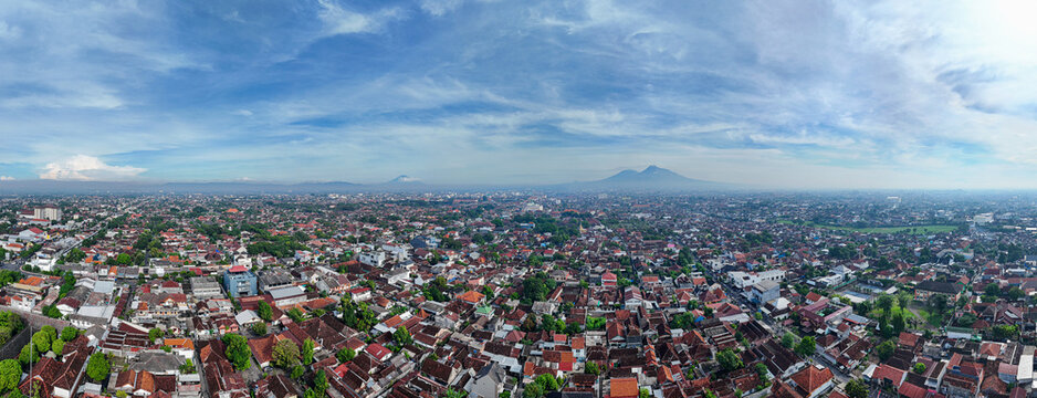 Aerial landscape panorama of Yogyakarta city at morning with Mount Merapi, Merbabu, and Sumbing volcanoes. Dense residential urban area and cityscape under a clear blue sky.