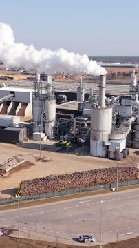 Vertical screen Wide Aerial Panorama Chimney Cloud Plume Over Production Plant With Stacked Timber, Fenced Road Frontage And Heavy Piping Framing The Manufacturing Complex
