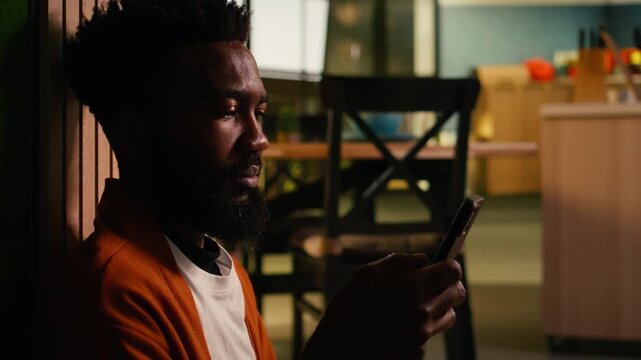 African American man using mobile phone indoors checking social media and texting in a cozy home environment. Warm interior and comfortable mood highlighting relaxation and free time