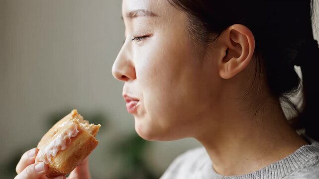 Asian woman deeply enjoying a glazed donut, savoring a sweet snack indoors.