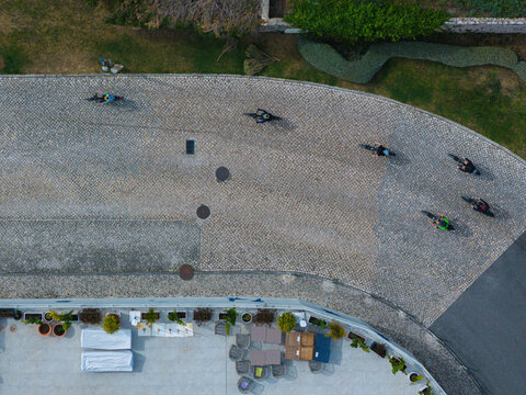 Aerial view of a group of cyclists riding on a curved cobblestone path next to a building terrace with outdoor seating, potted plants and green bushes under bright sunlight.