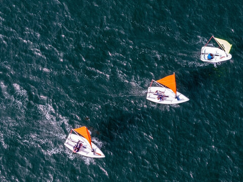 Aerial view of three small sailboats with orange and yellow sails sailing on deep blue water with people on board.