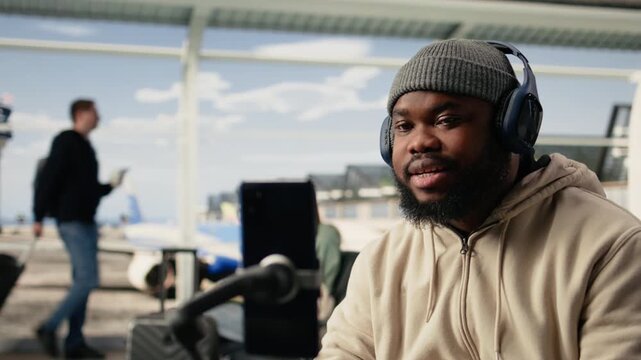 Black man vlogger capturing live broadcast on phone in airport terminal, documenting journey and holiday experience. Online streaming and modern mobile content creation by influencer.