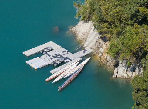Aerial view of a floating modular dock with long canoes and motorboats moored on turquoise water by a wooded bank in Fuxing District, Taoyuan City, Taiwan.
