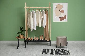 Naklejka premium Rack with stylish clothes, houseplant and pouf near green wall in room