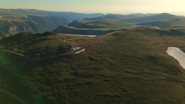 Beartooth Highway in Montana cuts through vast alpine terrain with snow patches and rolling green hills. Golden hour light illuminates the scenic mountain road stretching toward distant peaks.