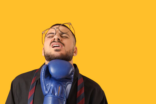 Young businessman getting hit in his face with boxing glove on yellow background. Knockout concept