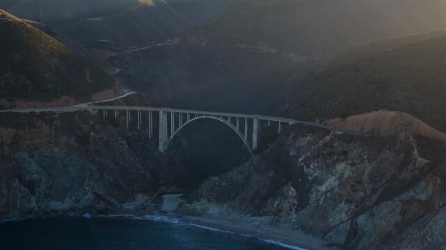 Aerial Bixby bridge over rugged coastline, sundrenched cliffs, arch span, crashing waves, mist rising, winding highway, cinematic golden light, drone perspective reveals scale, serene remote mood