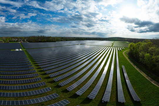 Aerial view of a large solar farm with rows of photovoltaic panels reflecting sunlight under a cloudy blue sky surrounded by green trees in Indianapolis, Indiana, United States.