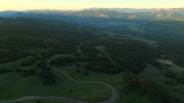 Aerial footage of Beartooth Highway in Montana winding through lush green valleys and dense pine forests. Distant mountain ranges glow under golden sunset light creating a breathtaking panorama.