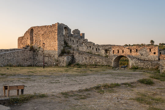 A wide landscape captures the impressive, crumbling stone walls of Berat Castle, surrounded by arid grass and featuring a simple wooden bench in Berat, Albania.