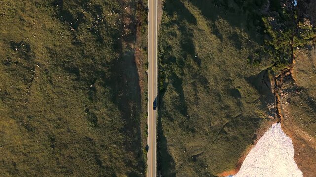 Drone footage follows a vehicle traveling along Beartooth Highway in Montana during sunset. Snow patches dot the rugged alpine terrain beside the winding mountain road.