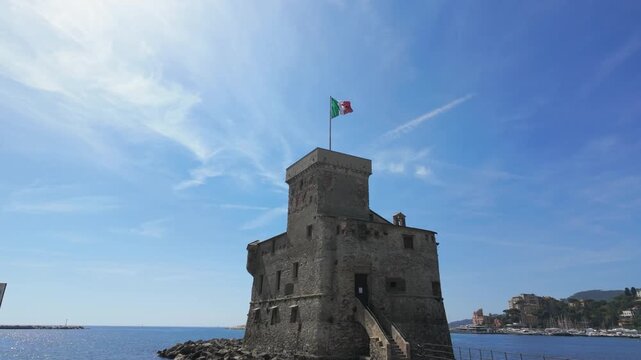 Drone video captures historic Rapallo Castle on the Ligurian coast, Italian flag fluttering atop stone tower, vivid blue sky, calm sea, and sunlit shoreline, no people visible, wide angle shot.