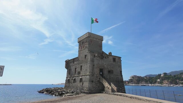 Historic Rapallo Castle with Italian flag stands on the Ligurian shoreline, surrounded by sparkling sea, vivid blue sky, and sunlight. Wide shot, no people, peaceful summer atmosphere.