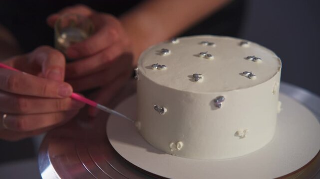 White cake decorated with silver embellishments. Hands carefully apply details with a pink tool. Person focused, meticulous, precise. Ideal for food, baking, or dessert projects