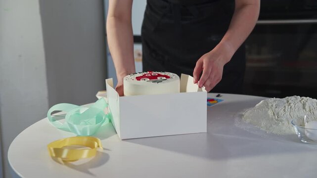 Person places cake into white box with care. Hands show gentle touch, focused attention. Table holds baking tools and flour. Ideal for food, baking, or gift presentation scenes