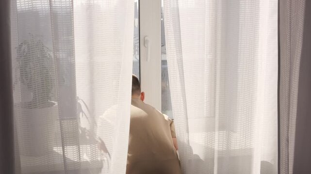 Man standing by window behind curtains at home window frames morning quiet solitude and reflection Window light creates soft mood isolation concept writer or freelancer lifestyle calm indoor scene