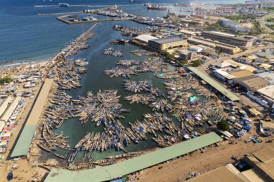 Aerial view of the bustling Tema Fishing Harbour with hundreds of traditional wooden canoes docked near industrial buildings and warehouses in Tema, Greater Accra Region, Ghana.