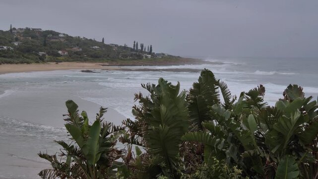 A view of a residential hillside, sloping down to a sandy beach with ocean waves rolling in under an overcast sky on the South Coast, KwaZulu-Natal, South Africa.